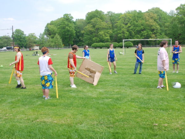 Eight athletes holding wiffle bats and cardboard appers on a soccer field.