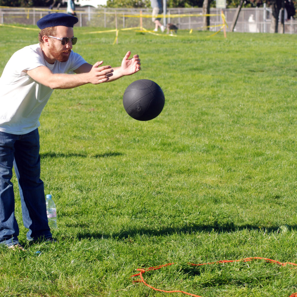 a man wearing beret hat and an avatar sunglasses throwing a black ball into a circle drawn in the field with a orange string.