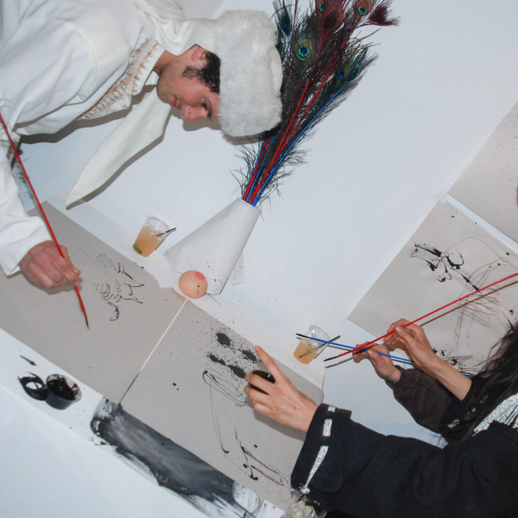 A man in a white dress and fur hat and a man with long straight hair, black dress and a sombrero drawing in ink with peacock weather pens.