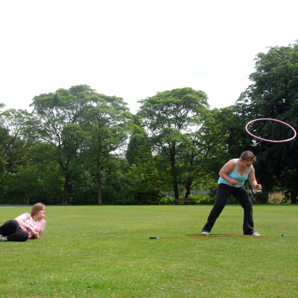 Two women sitting and watching another woman playing with a hoola hoop in open green field.