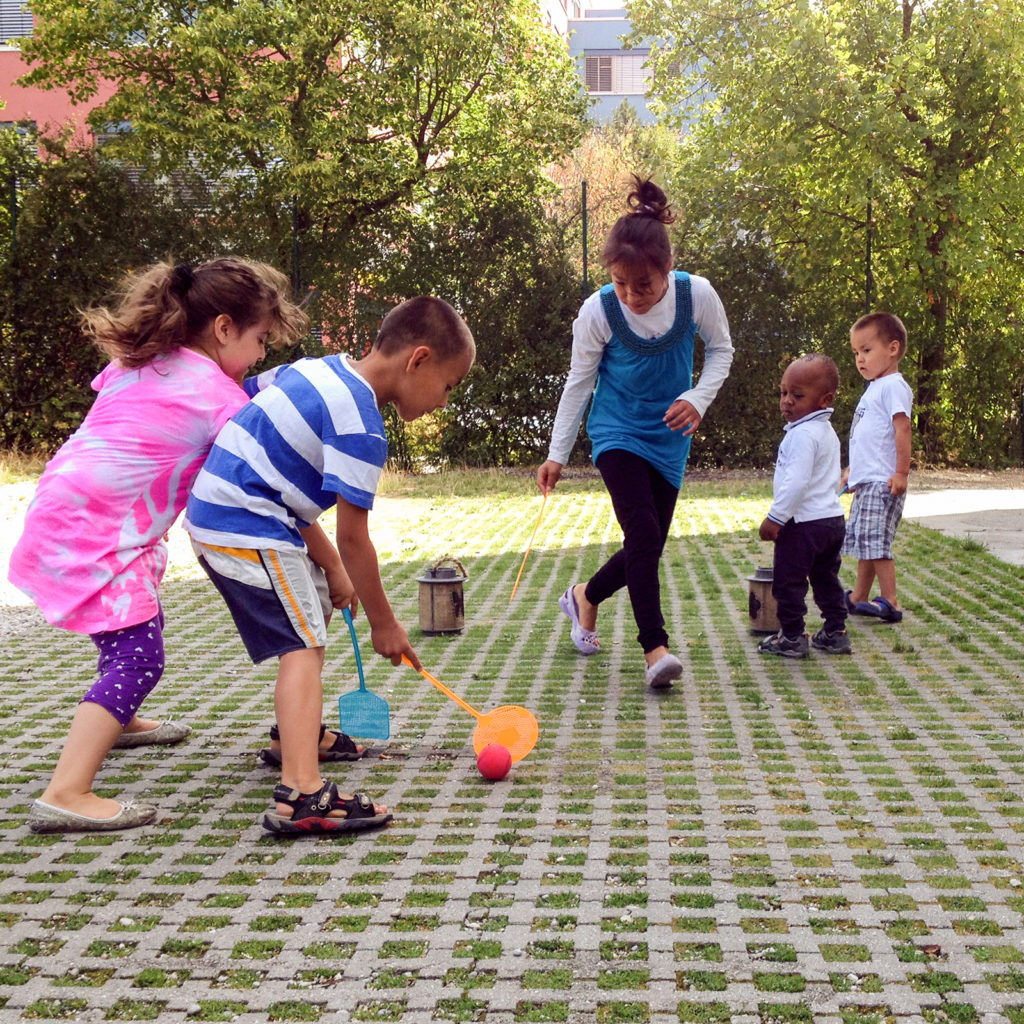 Five kids playing hockey like game with fly swatters and a red rubber ball outside.