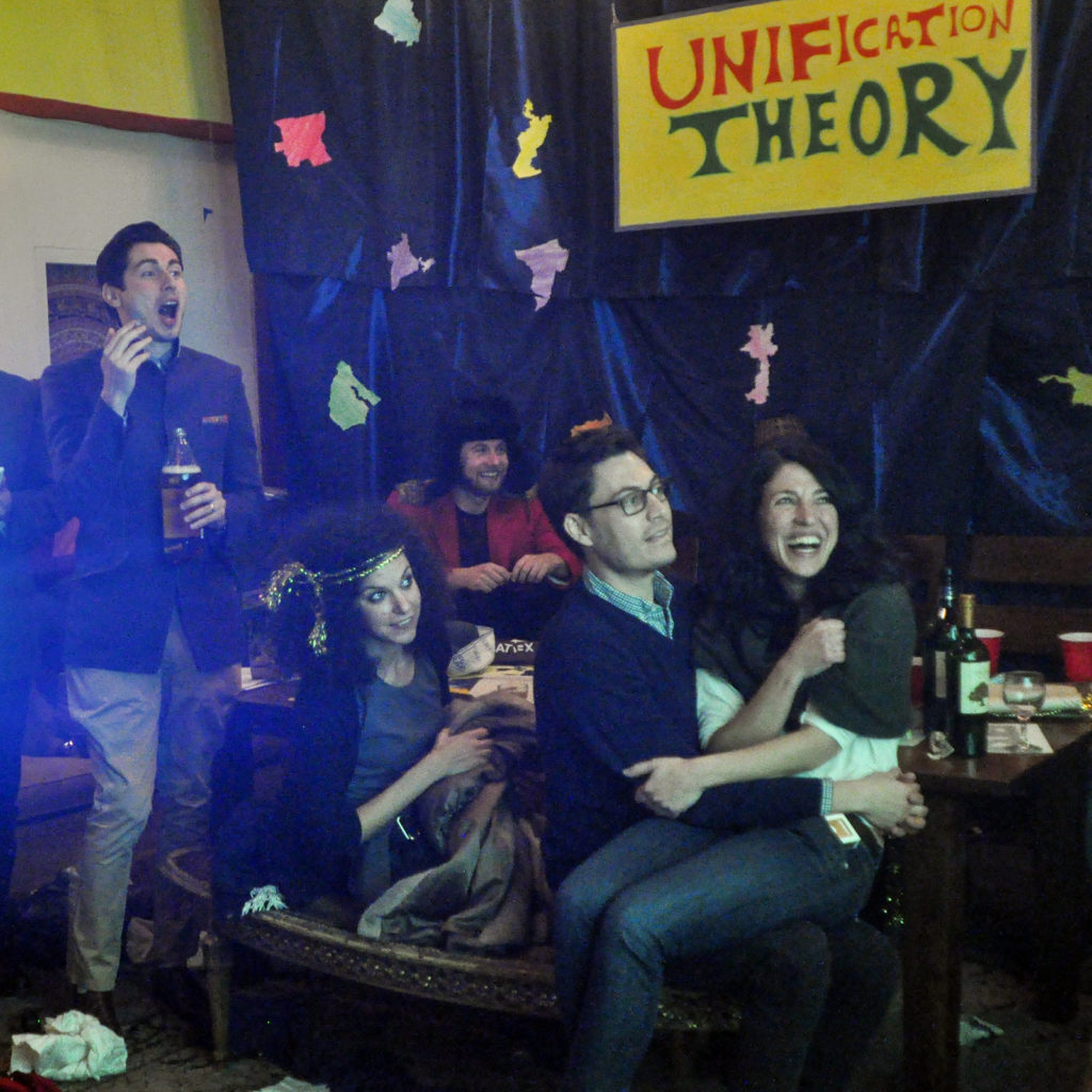 A crowd responding to what's being displayed on projector. A hand painted sign that reads "Unification Theory" is hanged in the back.
