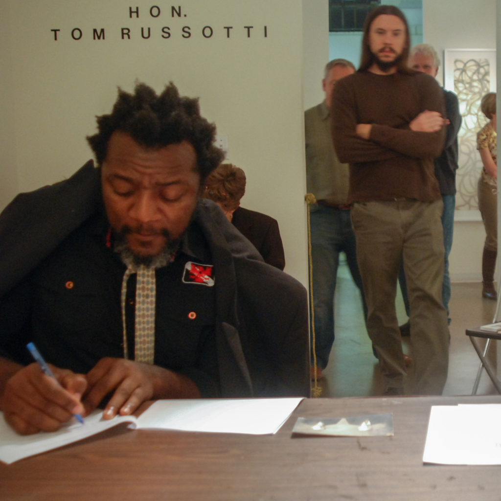 A man writing something on paper. He is being observed by visitors in the gallery. A text that reads "Hon. TOM RUSSOTTI" is seen on gallery wall in the back.