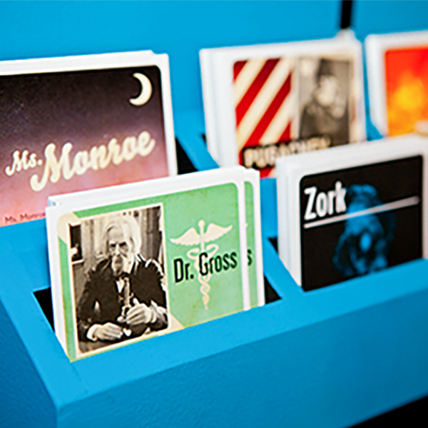 Six decks of cards in a sky blue display shelf. One deck reads, "Ms. Manrae," another reads, "Dr. Gross," and another one reads "Zork."