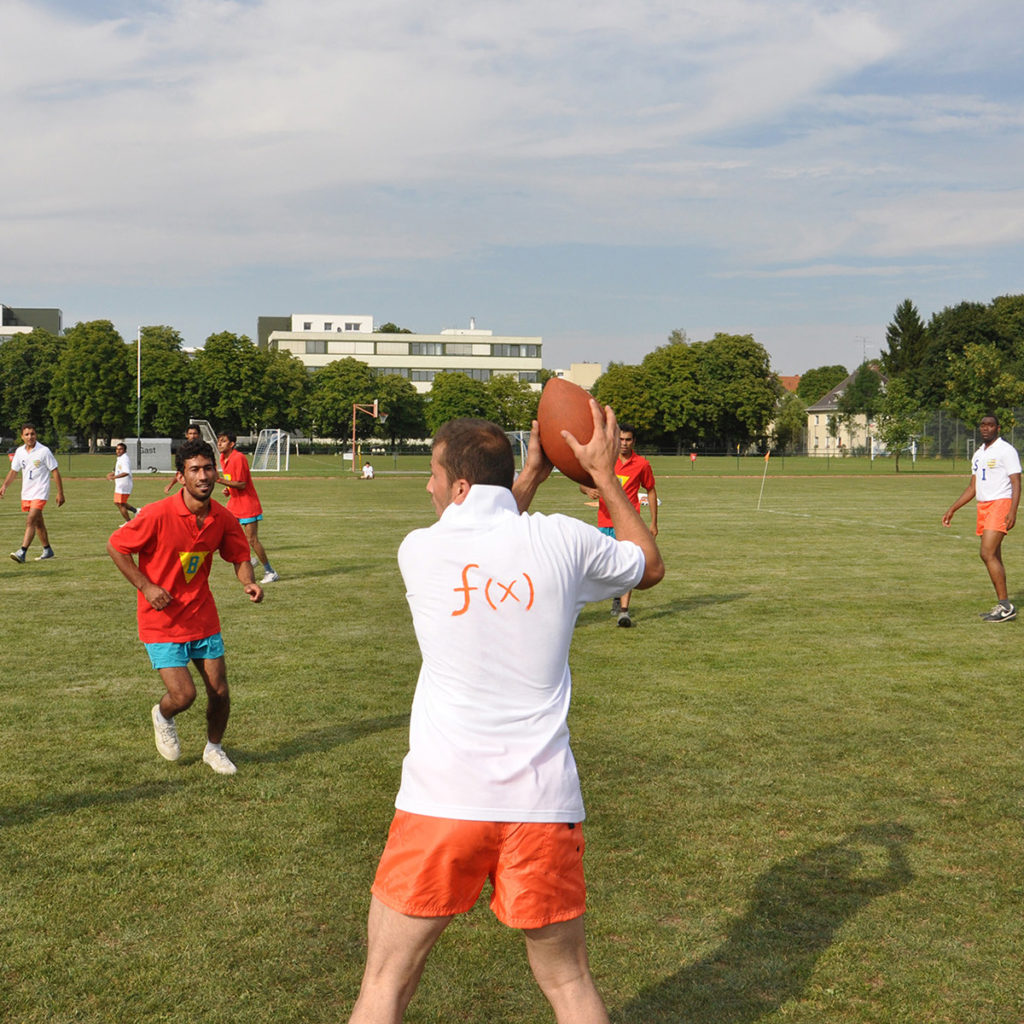 A man wearing a white shirt uniform with "f(x)" written on the back is about to throw an American football as another man with red shirt approaches him on a green field.
