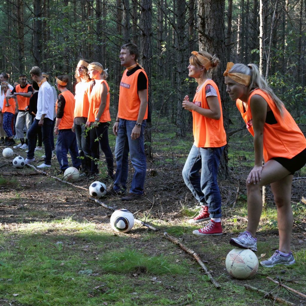 Over dozen men and women in orange shirt, each of them with a soccer ball, waiting for a kick off behind a line drawn with tree branches in a wood.
