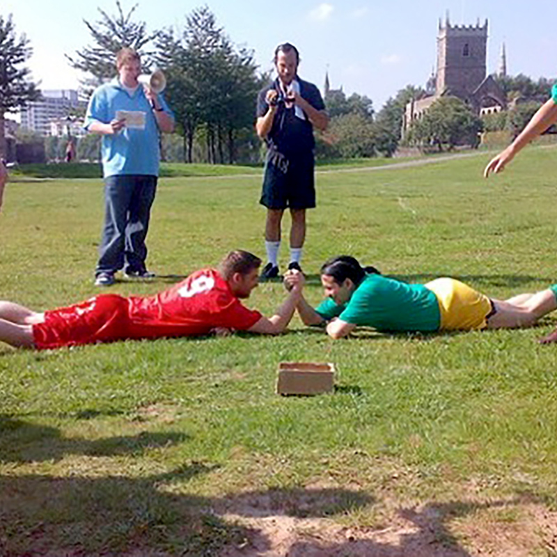 Two players, one in red uniform and another in green, arm wrestling on the ground surrounded by their teammates, an announcer and a man with video camera.