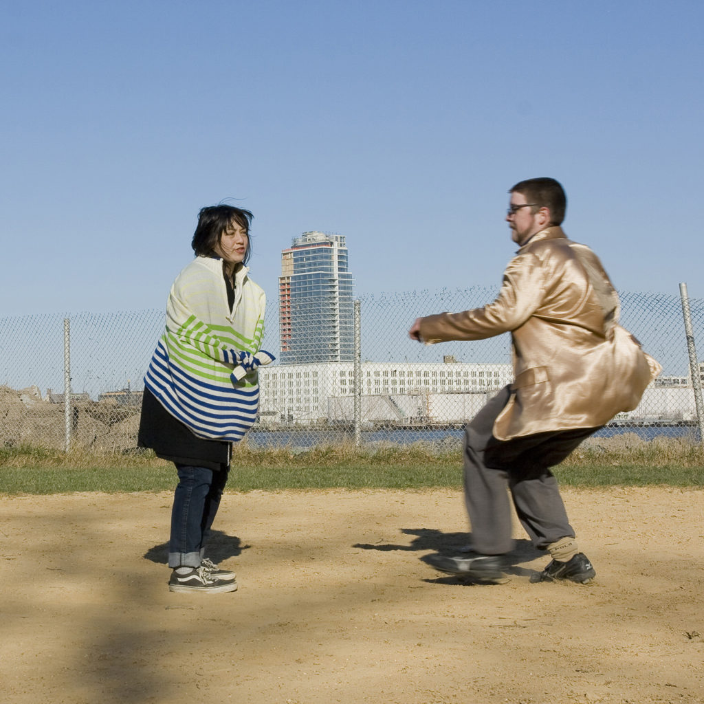 A woman in a straightjacket costume being chased away by a man in a gold jacket on a baseball field.