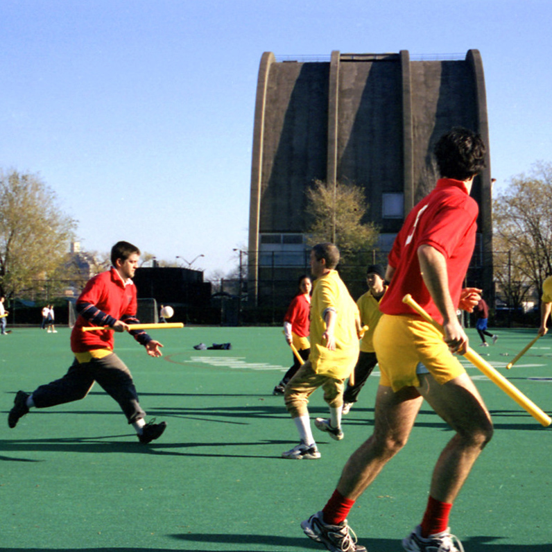 A man in red uniform runs across an artificial turf field full of other players in red yellow and red uniforms and holding wiffle bats as he bounces a wiffle ball with a waffle bat in the air.