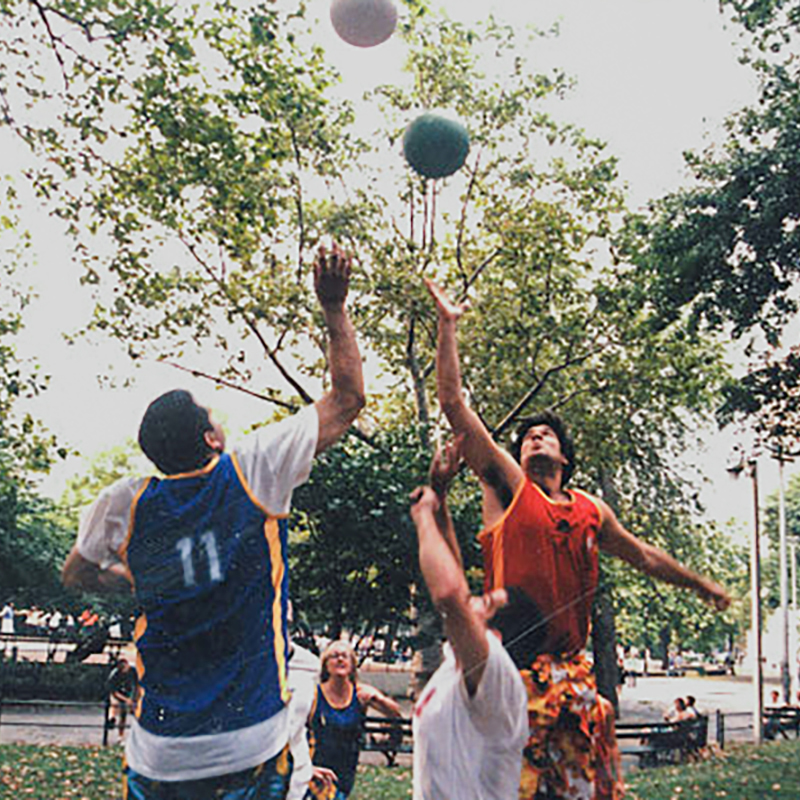 Two men are competing for a jump ball with two balls in the air on an outdoor basketball court.